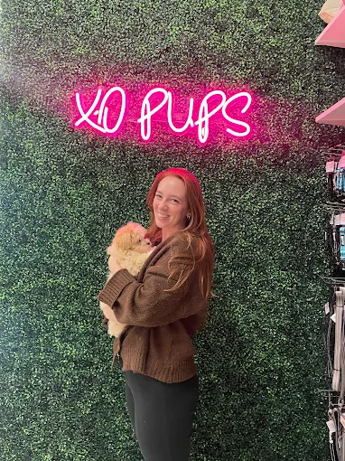 Woman holding a puppy in front of the illuminated XO Pups sign